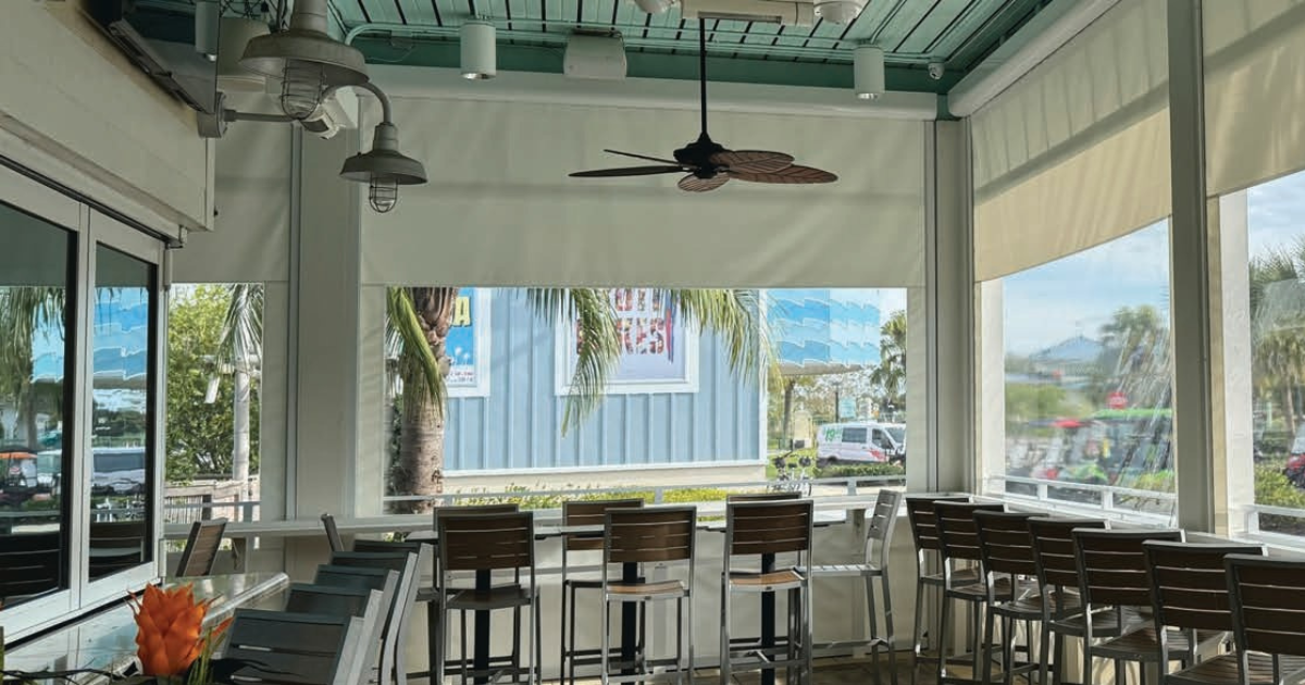 Restaurant patio featuring motorized clear panel enclosures with transparent weather protection panels, outdoor seating, ceiling fans, and clear visibility to the exterior.